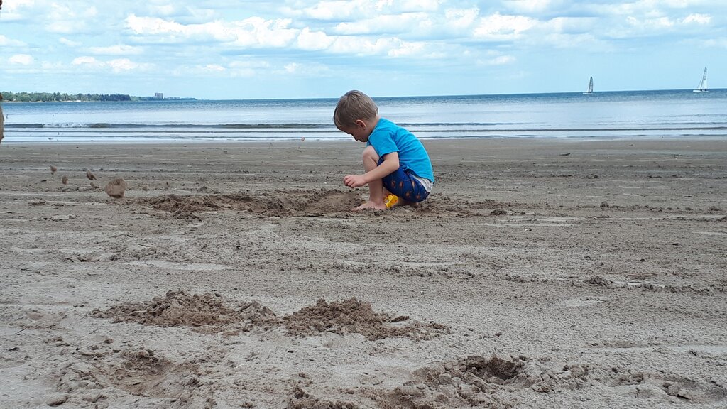 A Young child crouches on a sandy beach in Hamilton, digging and playing near the calm water. Sailboats are visible in the distance beneath a partly cloudy sky.