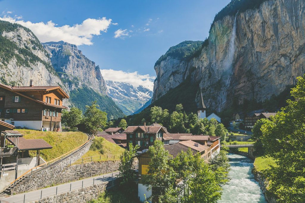 Alpine village in Switzerland nestled between steep rocky mountains under a clear blue sky. Wooden chalets and a white church sit beside a rushing turquoise river while tall waterfalls cascade down the cliffs.