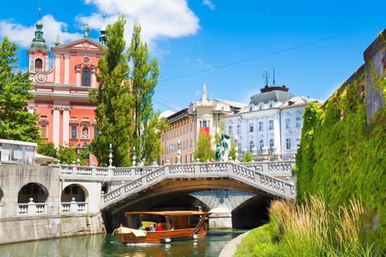 Wooden boat travels along a narrow river beneath an ornate stone bridge in a historic European city. Colorful architecture and lush green trees and ivy line the waterfront under a bright blue sky.