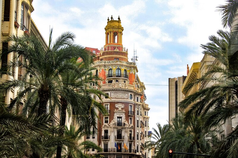 Palm trees line a city street with an ornate historic building rising between them. The building features decorative stonework balconies and a colourful round tower beneath a partly cloudy sky.