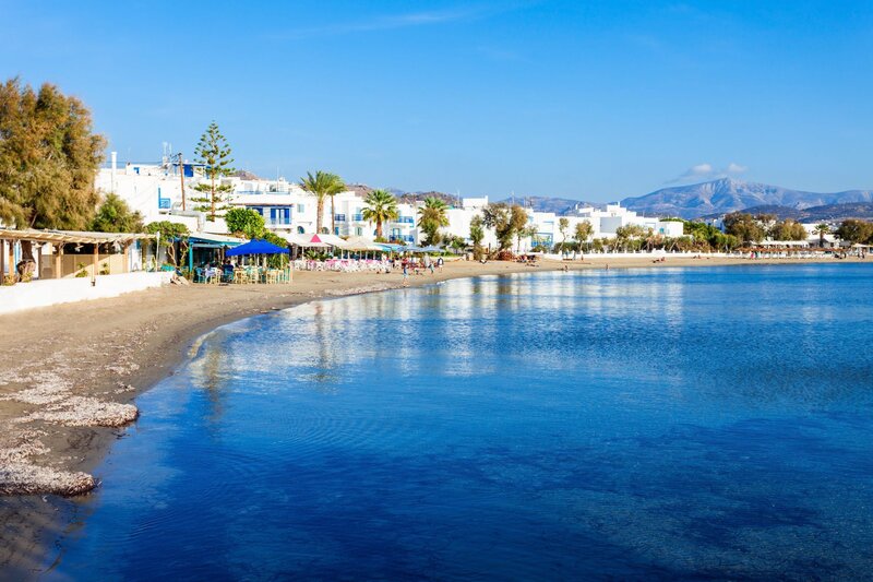 Curved sandy beach beside calm clear blue water with white buildings and palm trees lining the shore in Naxos, Greece. Mountains rise in the distance beneath a bright blue sky.