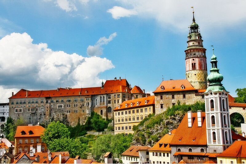 Historic castle complex with tall ornate towers and orange tiled roofs rising above a hillside town. Colorful old buildings and leafy green trees sit beneath a bright blue sky filled with large white clouds.