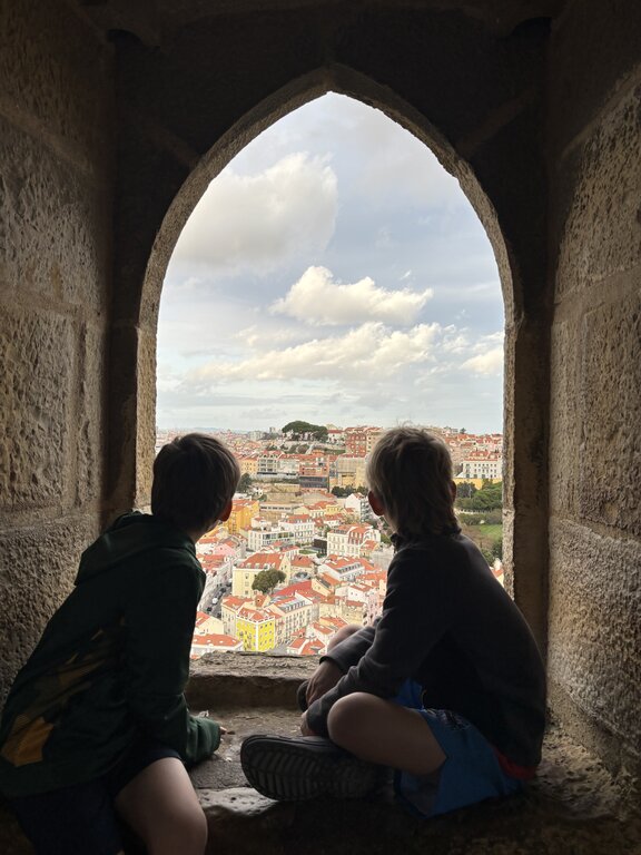 Two children sit inside a deep stone arched window and look out over Lisbon. Colourful buildings with red tiled roofs spread across the city below.