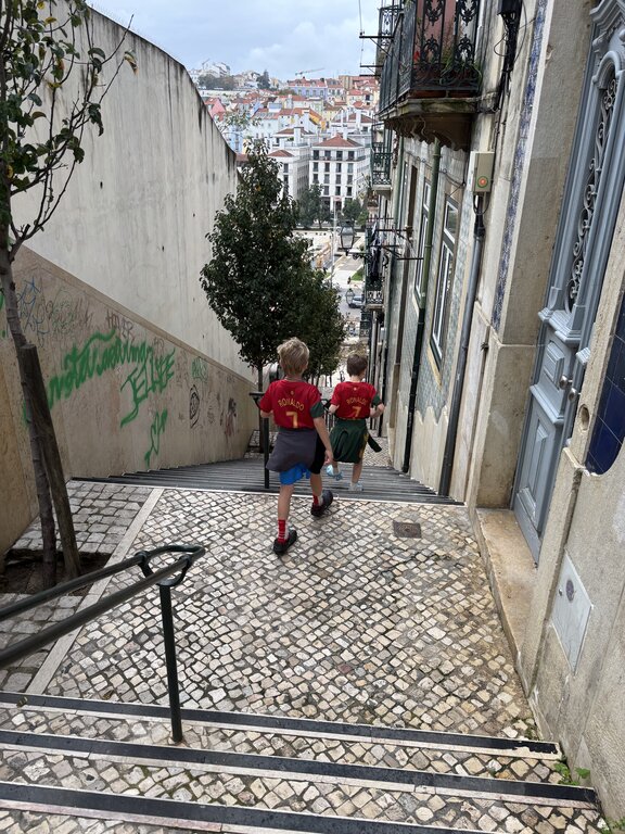 Two children wearing matching red Ronaldo 7 soccer jerseys walk down a steep cobblestone staircase in Lisbon Portugal between old buildings. Colourful hillside neighbourhoods fill the view ahead.