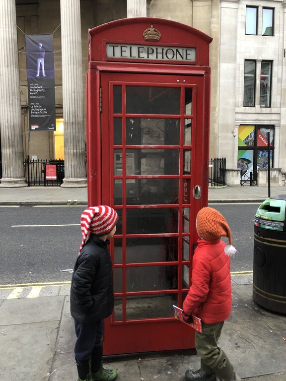 Two children in colorful winter hats stand beside a traditional red British telephone box on a London street. The top of the telephone box reads "TELEPHONE".