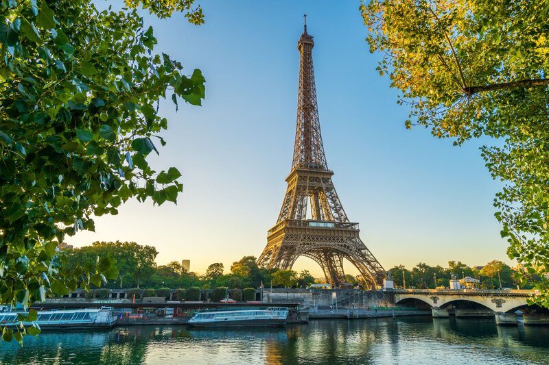 Eiffel Tower in Paris framed by leafy green trees in warm sunlight beside the Seine River. Sightseeing boats line the riverbank and a stone bridge crosses the water nearby.