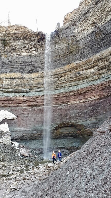 A narrow waterfall drops from the top of a layered rock cliff into a rocky basin below, where two hikers stand near its base. The exposed rock walls reveal distinct horizontal bands in shades of gray, brown, and red.
