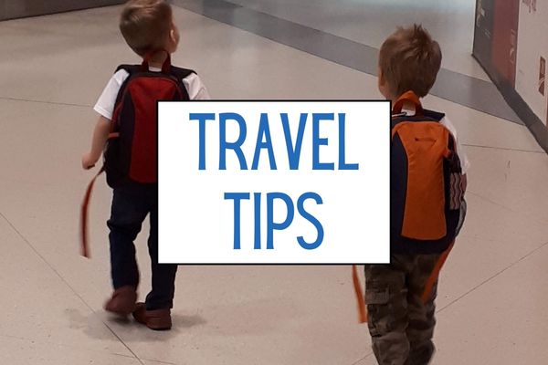 Two young children wearing backpacks walk side by side through an airport. A white graphic between them reads "TRAVEL TIPS".