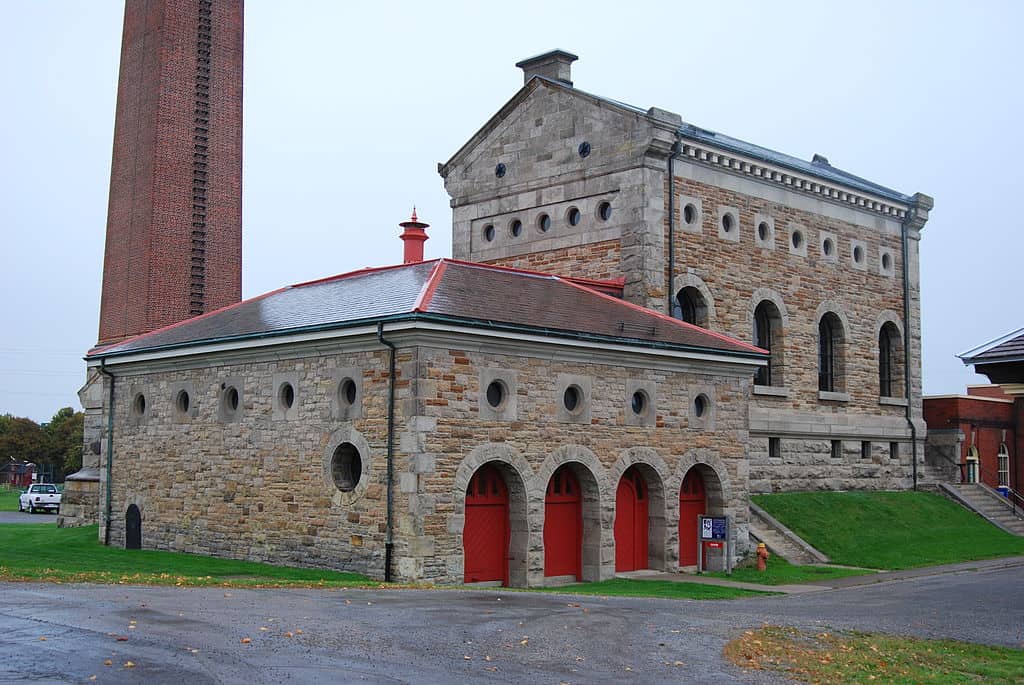 Historic stone industrial buildings with red doors and a tall brick smokestack at the Hamilton museum of Steam and Technology. The restored architecture sits on a grassy lawn under an overcast sky.