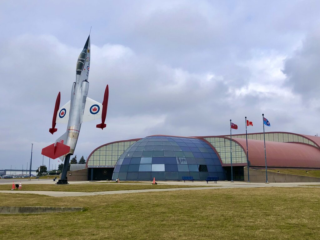 A Royal Canadian Air Force jet is mounted upright outside the Canadian Warplane Heritage Museum. The museum's curved glass entrance and three flagpoles stand beneath a cloudy sky.