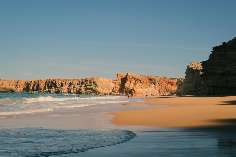Wide sandy beach in the Algarve Portugal, framed by rugged orange cliffs with gentle ocean waves rolling onto the shore. A few people swim and walk along the water in this scenic coastal destination for families visiting Europe.