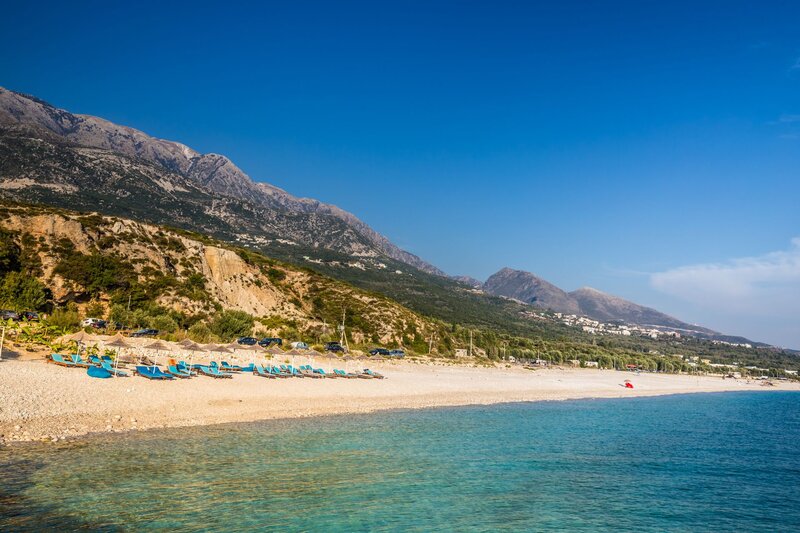 Long pebble beach lined with blue lounge chairs beside clear turquoise water in Dhermi, Albania. Steep rocky mountains covered in patches of green vegetation rise behind the quiet coastline under a cloudless blue sky.