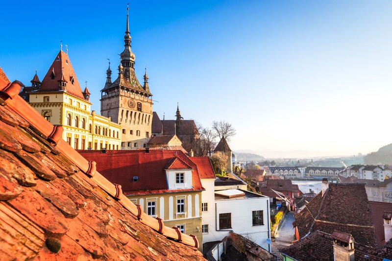 Historic town filled with red tiled rooftops and ornate medieval buildings glowing in warm sunlight. A tall stone clock tower with a decorative spire rises above the surrounding houses beneath a clear blue sky.