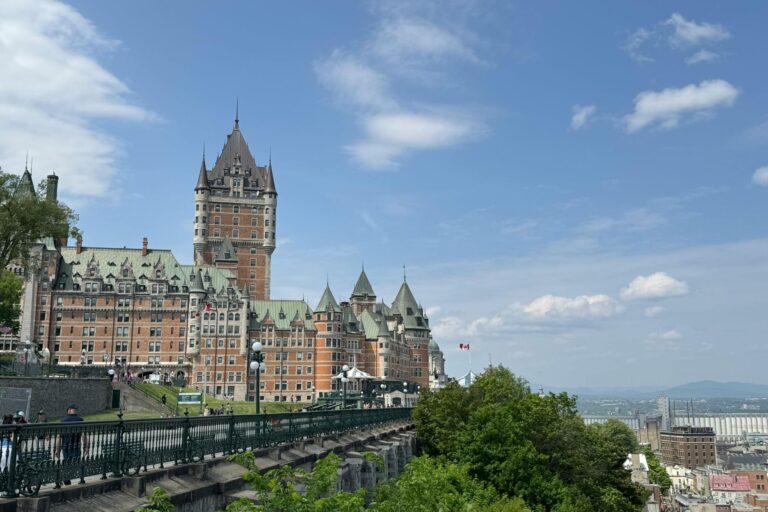 Château Frontenac rises above Québec City under a bright blue sky with scattered white clouds. The historic stone hotel with its copper green roofs overlooks a terrace lined with ornate railings, while trees and the city stretch into the distance below.