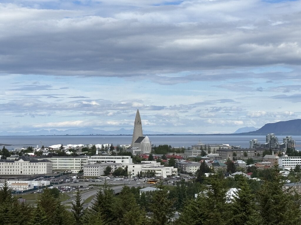 Reykjavik viewed from above with Hallgrímskirkja rising prominently among the low city buildings. Water and distant mountains stretch across the horizon beneath layered clouds.
