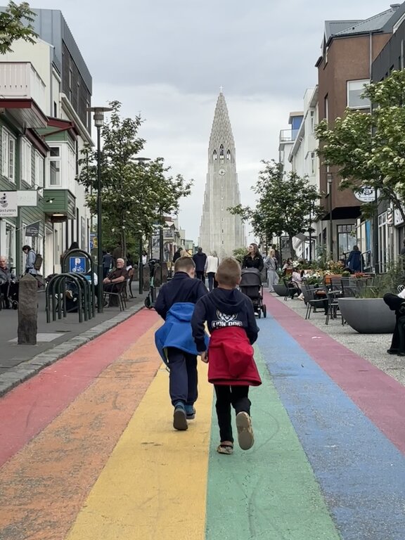 Two children walk along a rainbow painted street toward Hallgrímskirkja in Reykjavik. Cafes shops and pedestrians line the colourful street leading directly toward the tall church.