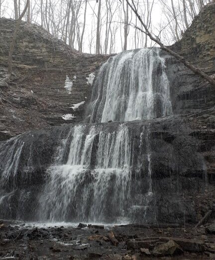 A two-tiered waterfall flows over dark rock ledges into a shallow rocky pool, with patches of lingering snow clinging to the cliffside. Bare trees rise above the gorge, giving the scene an early spring appearance.