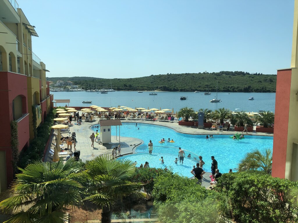 Families swim and play in a large outdoor pool surrounded by palm trees and rows of yellow umbrellas. Beyond the pool a calm blue bay is dotted with anchored sailboats and bordered by green hills.