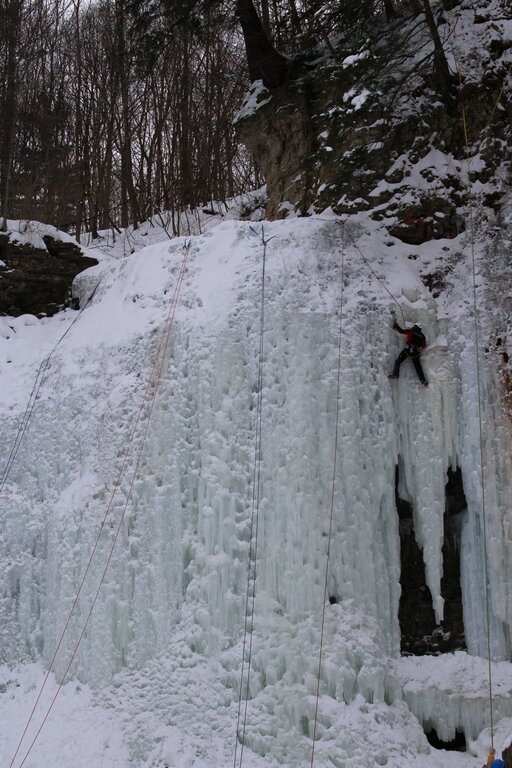 Ice climber scaling a massive frozen waterfall covered in thick icicles and snow. Climbing ropes hang down the icy cliff in the snowy winter landscape.