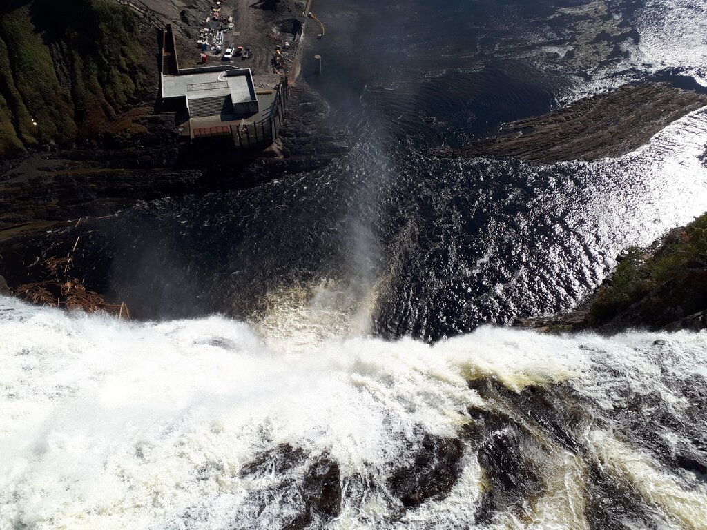 View looking straight down from the top of a massive waterfall as white water plunges over the rocky edge into the dark river far below. Mist rises from the base and emphasizes the dramatic height of the falls near Quebec City.
