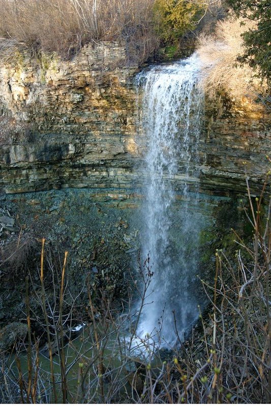 Wide waterfall pours over the edge of a dramatic rocky cliff into a deep gorge below. Bare branches frame the waterfall and exposed rock formations.