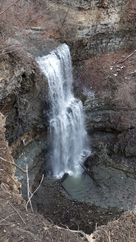 a Waterfall plunging over a rugged layered rock cliff into a small pool below. Bare trees and patches of snow surround the falls during the colder season.