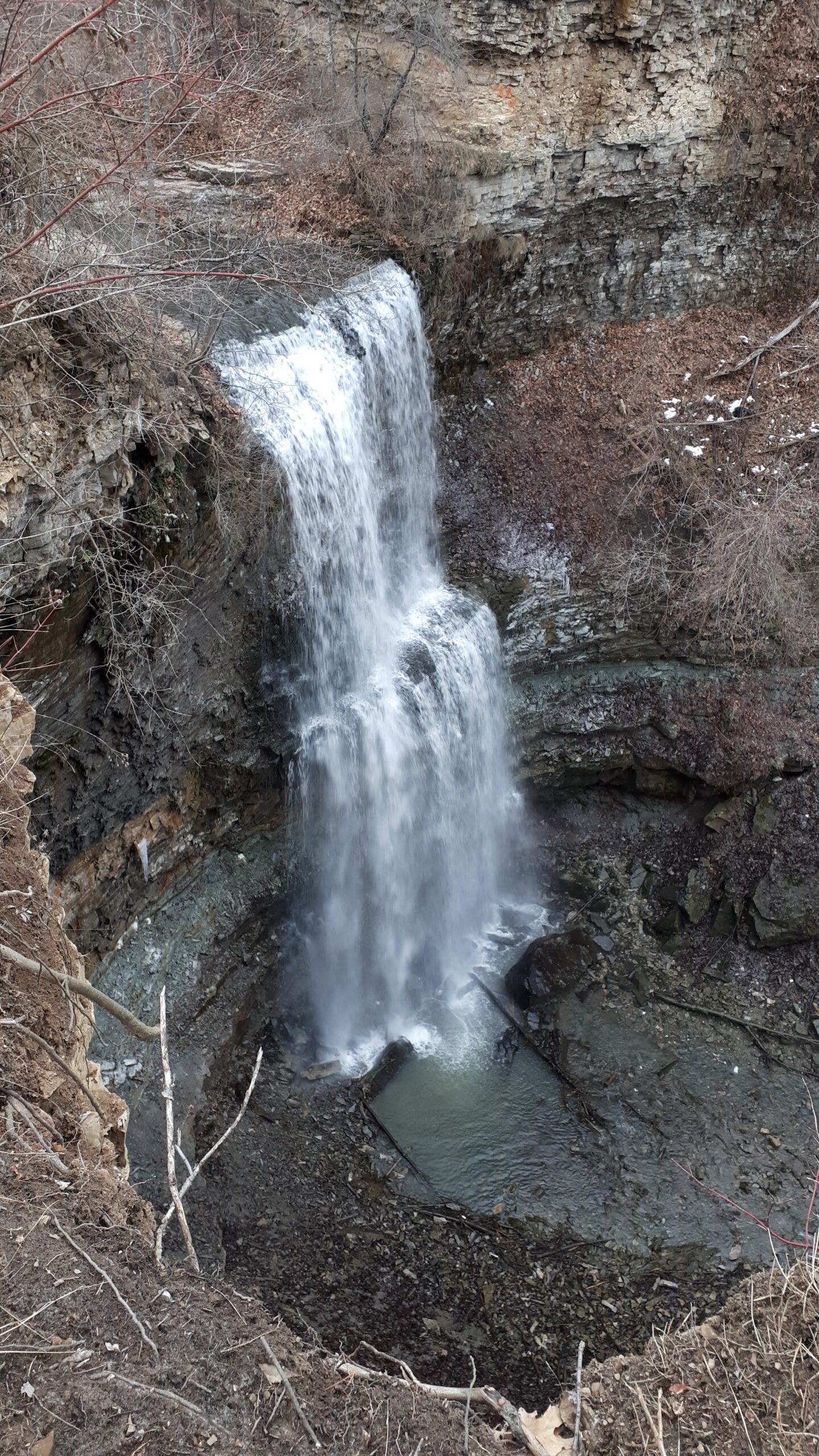 a Waterfall plunging over a rugged layered rock cliff into a small pool below. Bare trees and patches of snow surround the falls during the colder season.