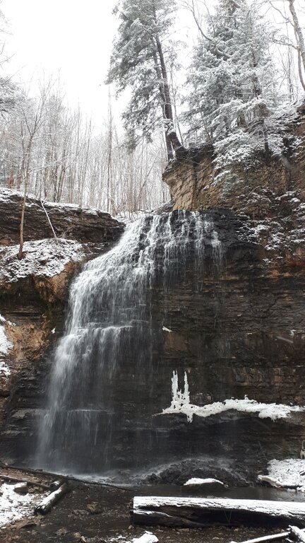 Waterfall in Hamilton Ontario cascading over a dark layered rock ledge in a snow covered forest in Hamilton Ontario. Snow dusts the surrounding cliffs evergreen trees and fallen logs.