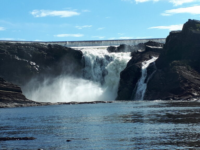Powerful waterfall near Quebec City rushing between dark rocky cliffs into a calm blue pool. A concrete dam stretches above the falls beneath a bright blue sky.