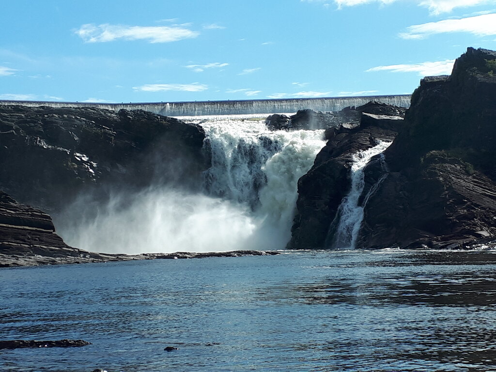 Powerful waterfall near Quebec City rushing between dark rocky cliffs into a calm blue pool. A concrete dam stretches above the falls beneath a bright blue sky.