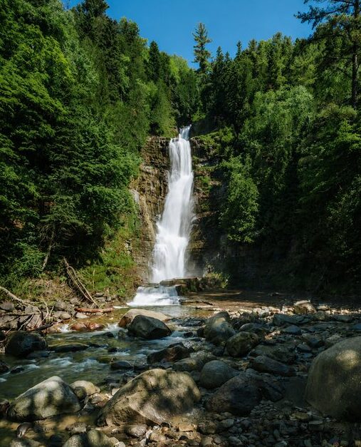 Chutes Jean Larose plunges down a tall rocky cliff surrounded by dense green forest near Quebec City. The waterfall flows into a shallow rocky creek scattered with large boulders.