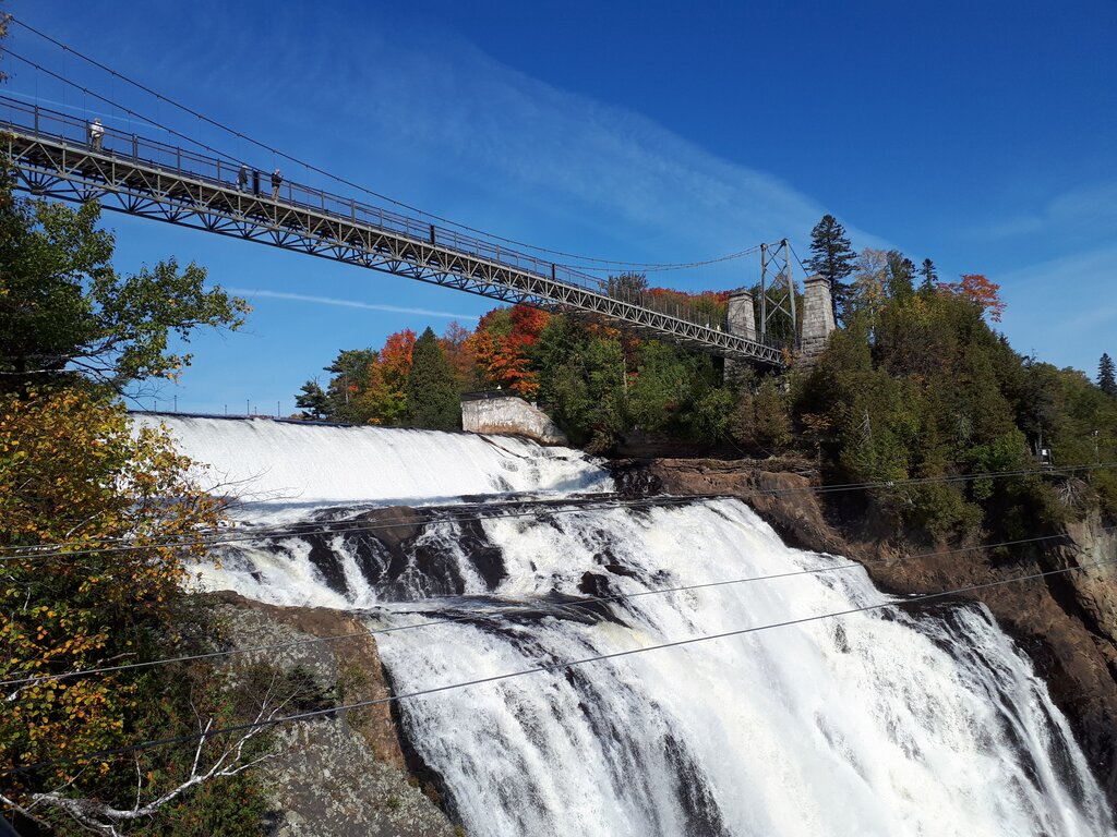 Powerful white waterfall cascades over a rocky cliff beneath a high suspension bridge. Brilliant red orange and green fall foliage surrounds the falls under a deep blue sky.