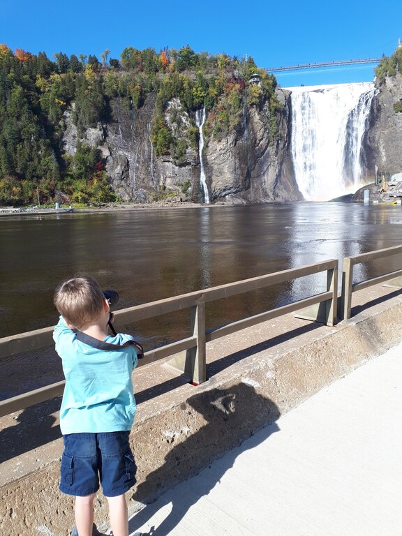 A child take a photo of Montmorency Falls, a towering waterfall near Quebec City surrounded by rocky cliffs and early fall foliage. A suspension bridge crosses high above the falls against a clear blue sky.