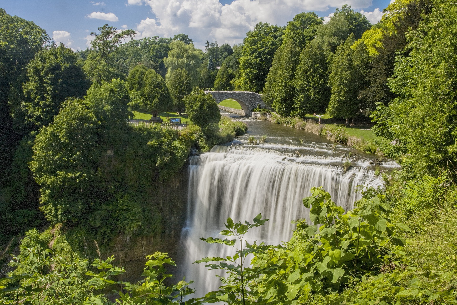 Wide waterfall cascading over the escarpment amid dense green summer foliage with a stone arch bridge upstream. The lush setting highlights the dramatic scenery found at waterfalls in Hamilton.