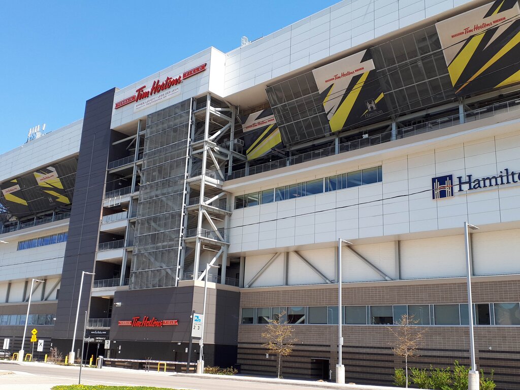 Exterior of Tim Hortons Field stadium in Hamilton Ontario beneath a clear blue sky. Large signs read "Tim Hortons Field" and "Hamilton".