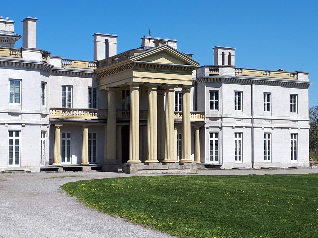 Large historic mansion with a pale stone exterior and tall classical columns beneath a clear blue sky. A curved driveway and green lawn lead to Dundurn Castle in Hamilton Ontario.