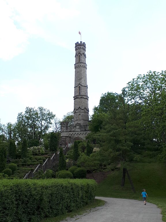 Tall stone tower rises above landscaped gardens and mature trees in a Hamilton Ontario park. A child walks along the path toward the tower and the long staircase leading up to it.