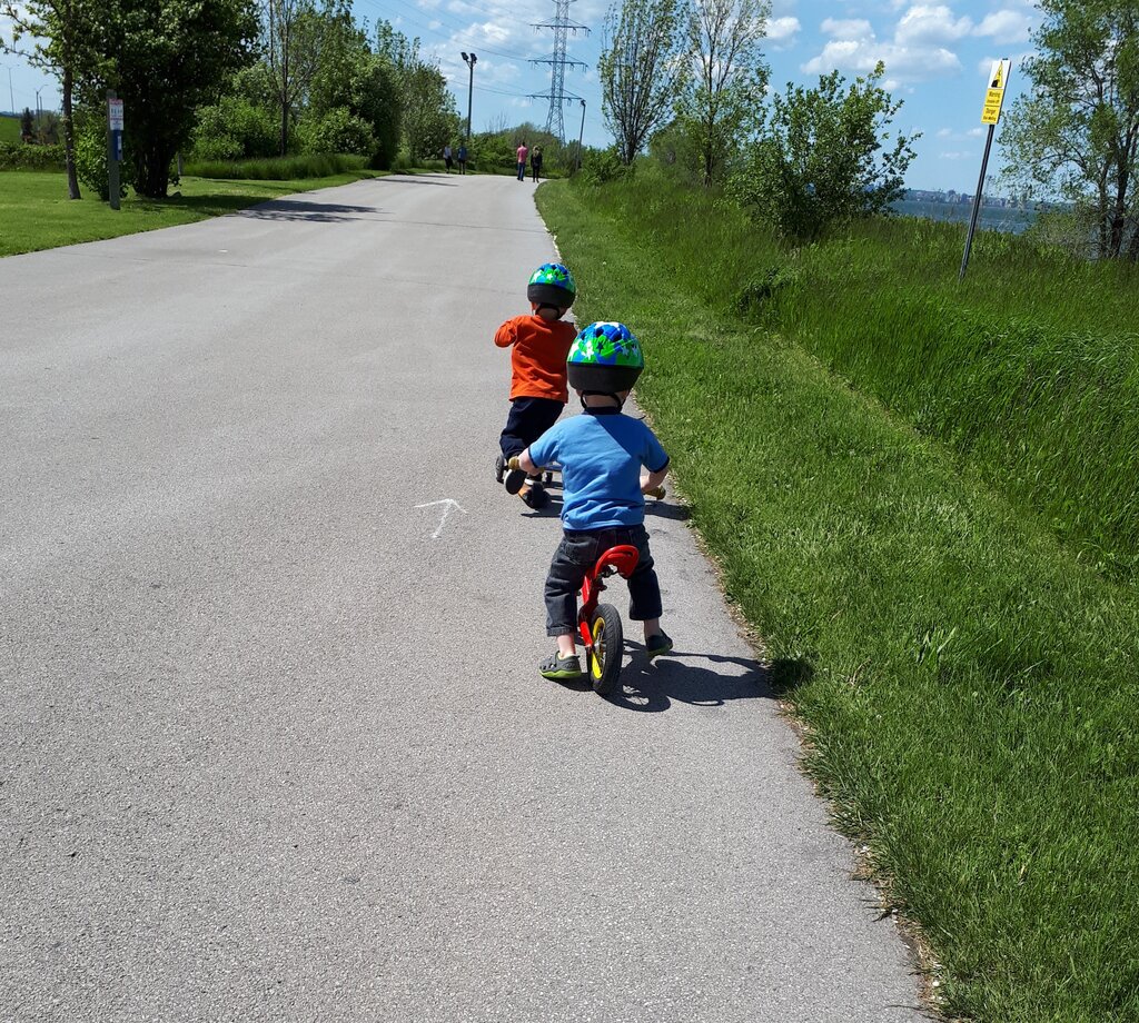 Two young children wearing helmets ride balance bikes along a wide paved waterfront trail in Hamilton Ontario. Green grass and trees line the path with water visible in the distance.