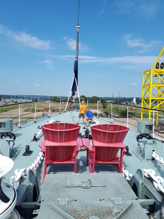 Two children explore the deck of a large ship tribal war ship (the Haida) with two bright red chairs overlooking Hamilton Harbour. Marina masts and industrial structures are visible in the distance beneath a blue sky.