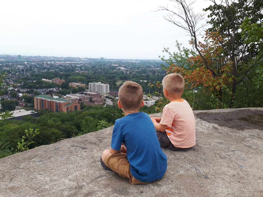 Two children sit together on a rocky lookout high above Hamilton Ontario. They face a broad view of neighbourhoods apartment buildings and dense green trees across the city.