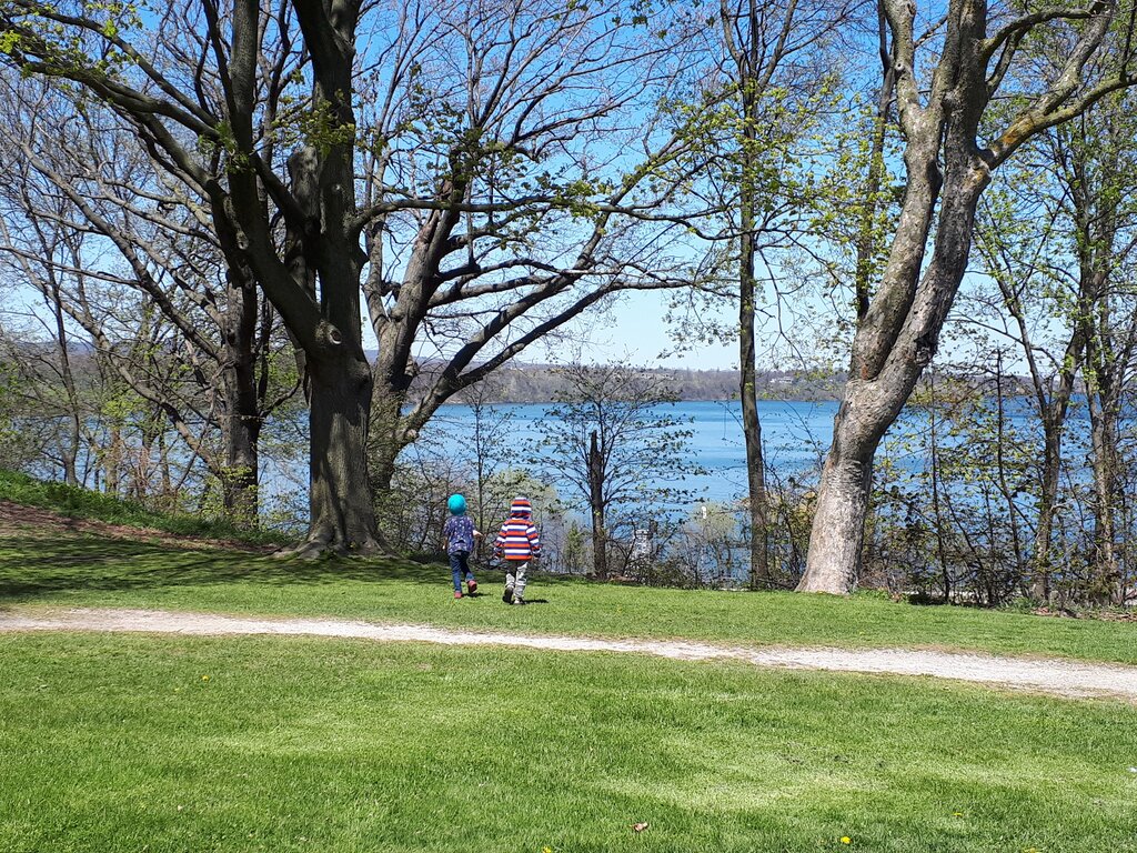 Two children walk across a green lawn toward the blue water beyond a row of large mature trees. The sunny park in Hamilton Ontario is beginning to turn green in spring and gives a good view of the water opposite Dundurn Castle