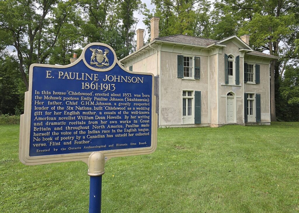 Chiefswood National Historic Site with the historic childhood home of Mohawk poet E. Pauline Johnson behind a provincial plaque describing her life from 1861 to 1913. The cream coloured house has green shutters and sits on a grassy property surrounded by mature trees.