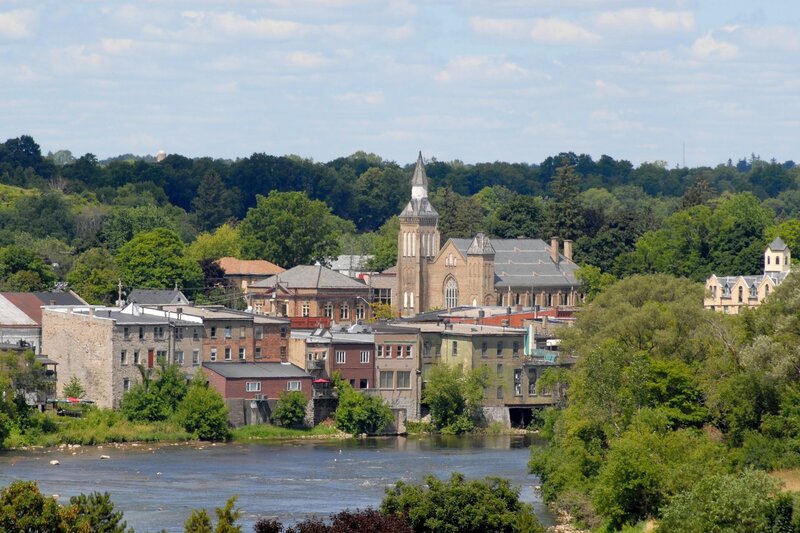 A small Ontario town surrounded by lush green trees with a river in the foreground and a historic stone church rising above downtown. The peaceful waterfront scenery makes this a picturesque option for day trips from Hamilton.