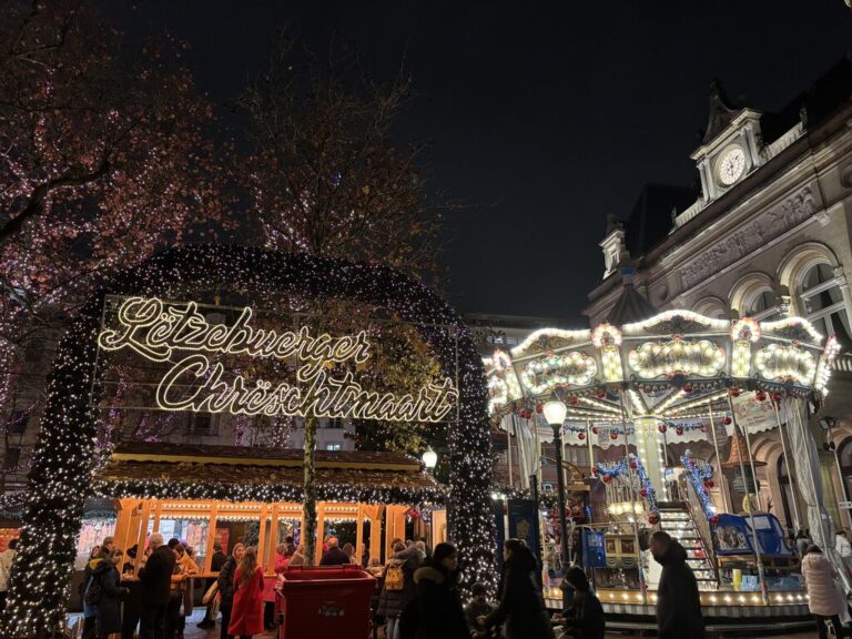 The Luxembourg Christmas market at night with a glowing sign reading "Lëtzebuerger Chrëschtmaart" beneath an arch covered in white lights. A brightly illuminated carousel and wooden market stalls are surrounded by visitors.