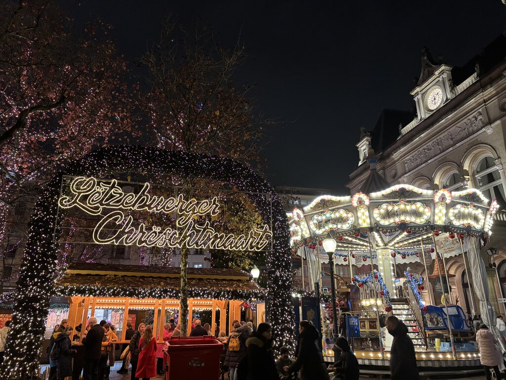 The Luxembourg Christmas market at night with a glowing sign reading "Lëtzebuerger Chrëschtmaart" beneath an arch covered in white lights. A brightly illuminated carousel and wooden market stalls are surrounded by visitors.