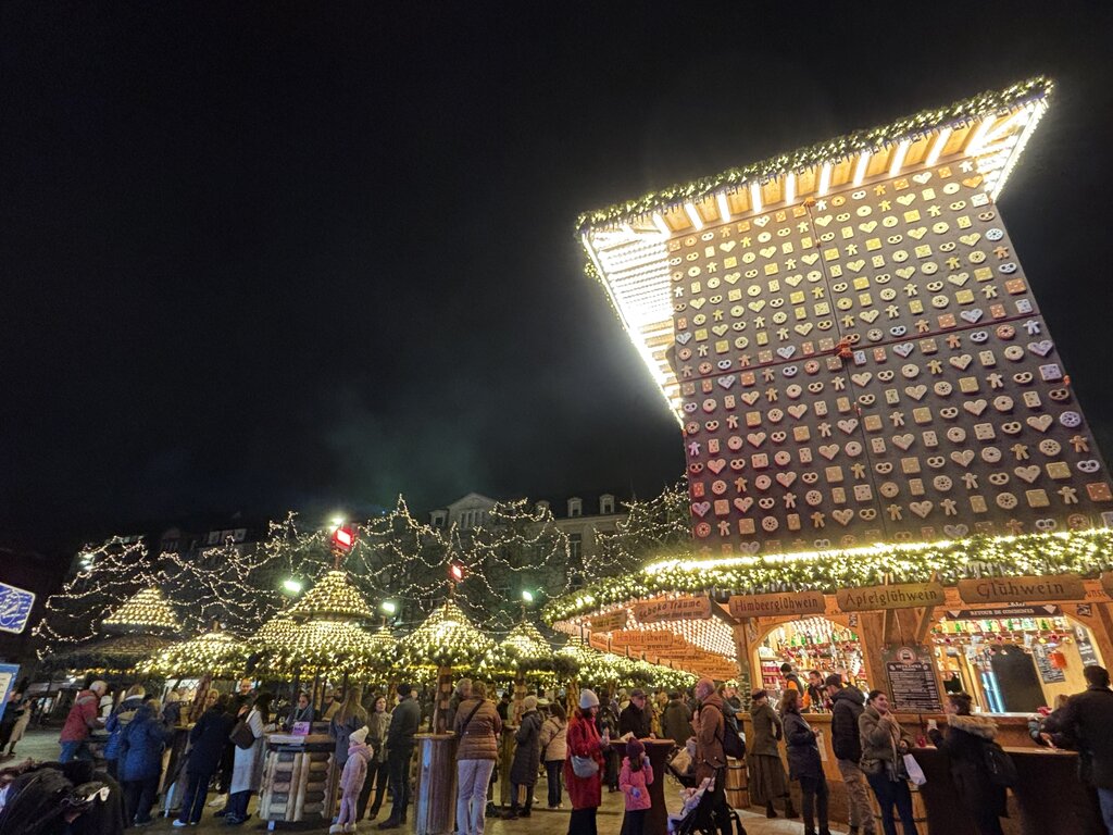 Visitors gather around festive drink stalls beneath a towering gingerbread themed Advent calendar at a Luxembourg Christmas market. Signs on the wooden stall read "Himbeerglühwein" "Apfelglühwein" and "Glühwein".
