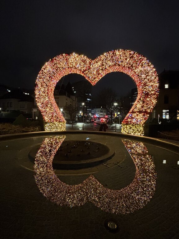 A giant heart shaped arch covered in red and golden Christmas lights stands over a fountain in Luxembourg at night. The glowing heart is reflected almost perfectly in the dark water below.