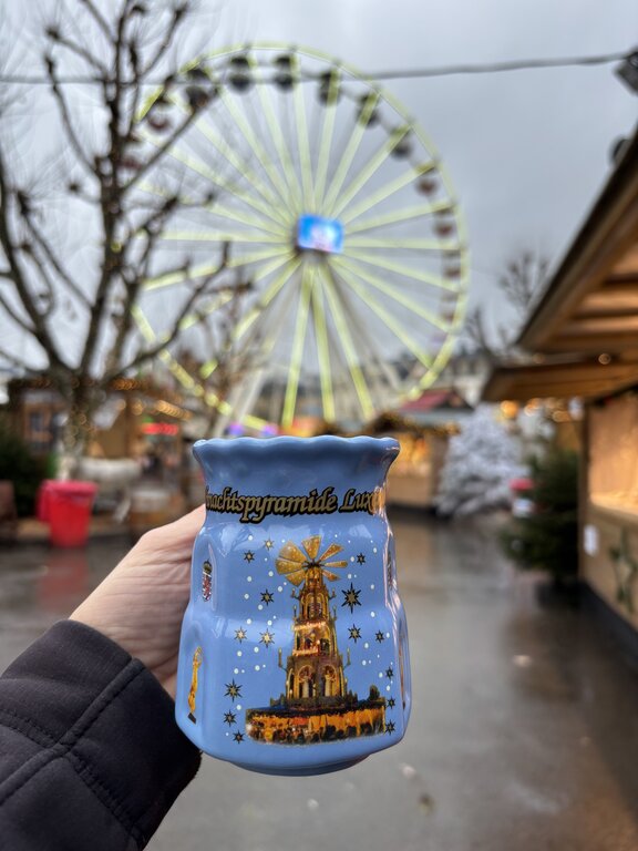 A hand holds a blue Christmas market mug decorated with a Christmas pyramid in front of a Ferris wheel in Luxembourg. Wooden stalls and holiday decorations fill the market in the background.