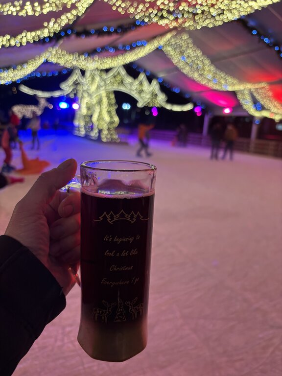 A hand holds a tall cup of hot Christmas market drink beside an outdoor ice skating rink in Luxembourg. Cascades of warm white lights hang overhead while skaters move across the colourful rink.