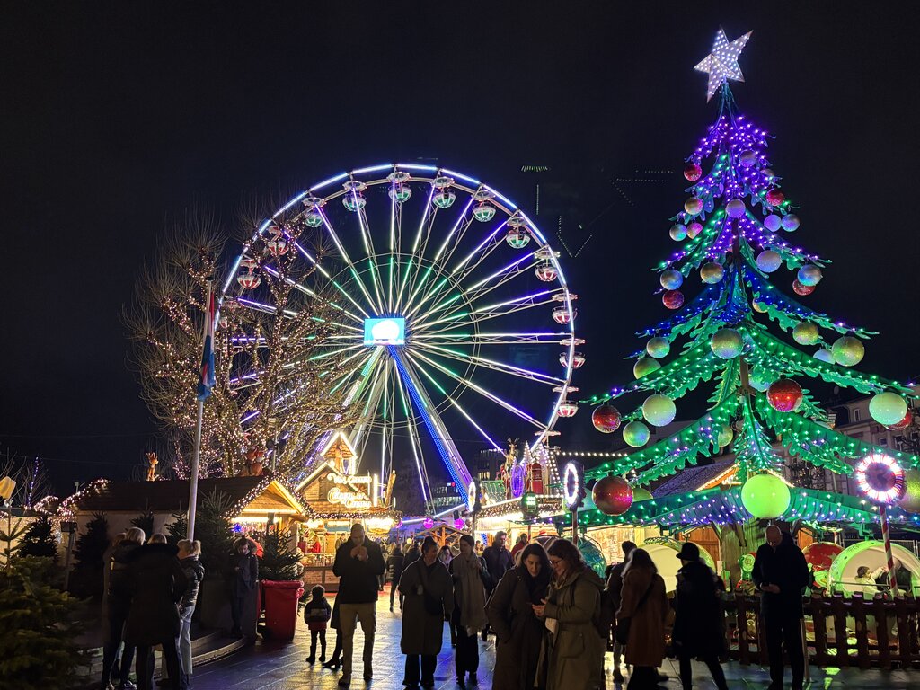 A Luxembourg Christmas market at night with a brightly lit Ferris wheel beside a huge Christmas tree covered in colourful lights and ornaments. Visitors walk between festive market stalls beneath the attractions.